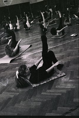 Aerial shot of a 1970s-era indoor yoga/stretching class in a large, wood-floored gym. Participants perform floor exercises on...