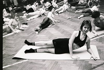 Group fitness class in a gymnasium, likely 1970s–1980s. Women in form-fitting athletic wear (leggings, tank tops) perform flo...