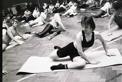 Group fitness class in a gymnasium, likely 1970s–1980s. Women in form-fitting exercise attire perform floor exercises on mats...