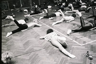 Group fitness class in a gymnasium, likely 1960s–1970s. Women performing floor exercises in coordinated poses, wearing form-f...