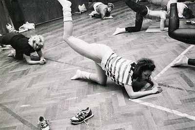 1970s indoor fitness class with participants in dynamic floor exercises, likely Pilates or yoga. Wooden flooring and casual a...