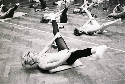 Group fitness class in a gymnasium, 1970s. Women perform floor exercises on mats in a large, wood-floored hall. Foreground su...