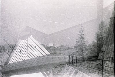 Vintage alpine lodge framed by snow-laden pine trees, set against misty mountain peaks. Geometric, pyramid-roofed structure w...