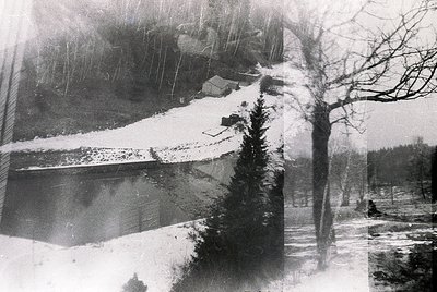 Vintage black-and-white winter scene featuring a snow-covered alpine lake bordered by bare trees and rocky terrain. Heavy sno...