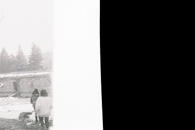 Black-and-white winter scene featuring three figures in heavy coats near a snow-covered structure. Dense fog obscures distant...