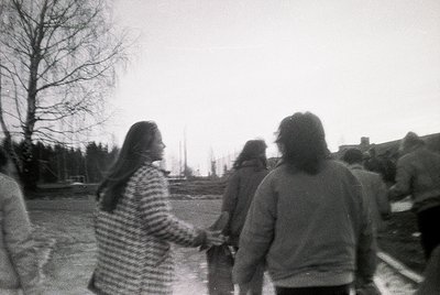 Black-and-white candid of three figures walking outdoors, holding hands. The woman in striped sweater leads, with blurred urb...