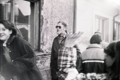Black-and-white street scene featuring a central figure in a plaid shirt and sunglasses gesturing with arms outstretched. Sur...