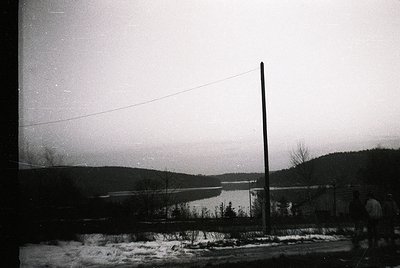 Vintage black-and-white landscape featuring a frozen lake bordered by leafless trees and rolling hills. A lone utility pole a...