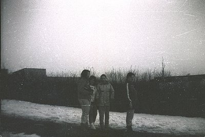 Vintage black-and-white photo of four individuals standing outdoors in winter, posing on snow-covered ground. The central fig...