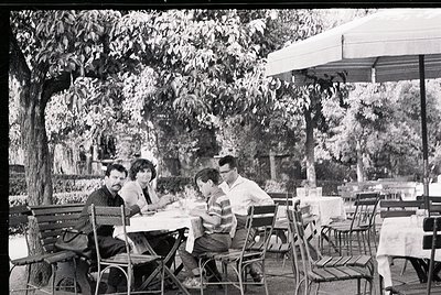 Mid-century outdoor café scene with four seated patrons at metal-framed tables under large umbrellas. Lush greenery and matur...