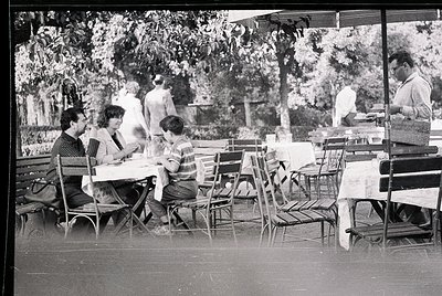 Mid-century outdoor café scene with metal-framed chairs and tables under leafy trees. Three adults and a child seated, engage...