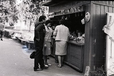 Street-side kiosk with vintage 1960s-70s design, featuring shelves stocked with jars, bottles, and packaged goods. Three peop...