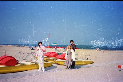 Vintage beach scene with two women in 1970s swimwear posing near yellow kayaks and red beach umbrellas. Clear blue sea and sa...