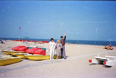Three individuals pose on a sandy beach with inflatable boats and umbrellas in background. Bright summer attire suggests mid-...