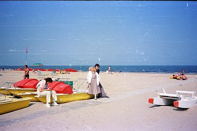 Vintage seaside scene with mid-20th century beachgoers. Yellow rowboats, red umbrellas, and a lifeguard tower frame a sandy s...