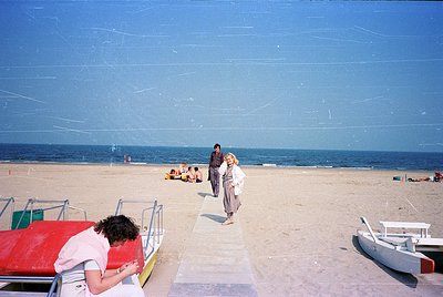 Vintage seaside scene with a sandy beach and shallow waters. A woman in a white dress sits on a red-and-white striped deck ch...