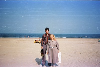 Two individuals pose on a sandy beach under clear skies, likely mid-20th century. The man wears a striped sweater and pattern...
