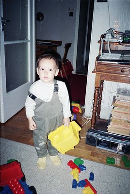 Toddler in 1970s-style overalls and white long-sleeve shirt stands indoors, holding a yellow cloth. Surrounding toys include ...