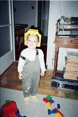 Young child wearing a yellow Lego headpiece and overalls, standing on wooden floor in a mid-century home. Surrounding toys in...