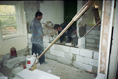 Two construction workers in a mid-renovation interior, likely residential. One stands holding a trowel, wearing a patterned s...