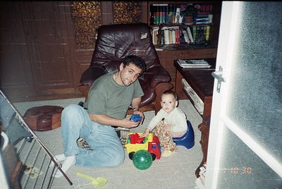 A man and child play with colorful plastic toys on a carpeted floor in a mid-century interior. Wooden bookshelves filled with...
