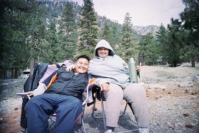 Two individuals pose outdoors in a forested area, likely during the 1990s–2000s. One wears a backpack, the other a vintage-st...
