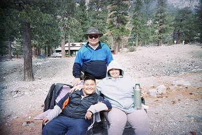 Three individuals pose outdoors in a forested area, likely a national park or campground. The man stands in a teal jacket and...