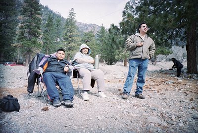 Three individuals in outdoor mountain setting, likely 2000s. Man in foreground wears beige jacket, jeans, and hiking boots; s...
