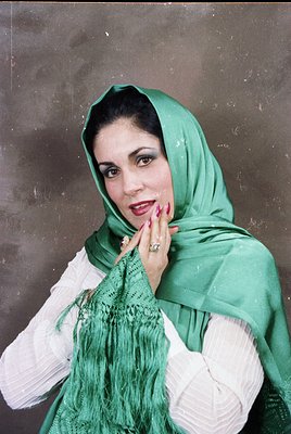 Portrait of a woman in traditional Middle Eastern attire, featuring a green headscarf and white embroidered garment. Her pose...
