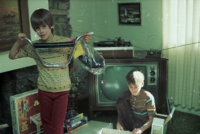 Vintage indoor scene featuring two boys in 1970s-style striped sweaters and red pants. One boy holds a transparent plastic ba...