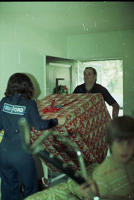 Vintage indoor scene featuring two men handling a large, patterned red-and-white fabric (likely a bedspread or curtain) in a ...