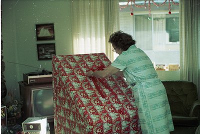 Mid-century living room interior featuring a woman in a light-colored dress dusting a floral-patterned sofa (red/cream). Blac...