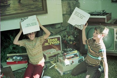 Two boys in 1970s-style striped shirts balance cardboard boxes on their heads indoors, likely a 1970s living room. Boxes labe...