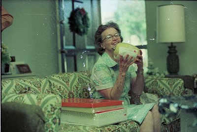 Vintage indoor scene: woman in 1970s-style floral dress drinks from a yellow ceramic cup, seated on patterned sofa. Side tabl...
