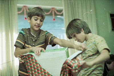Two boys in 1970s-style striped shirts and plaid aprons play with a toy train set indoors, near a window adorned with festive...
