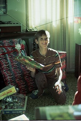Vintage photo of a boy in the 1970s holding a colorful, retro surfboard in a cluttered indoor setting. Surrounding items incl...