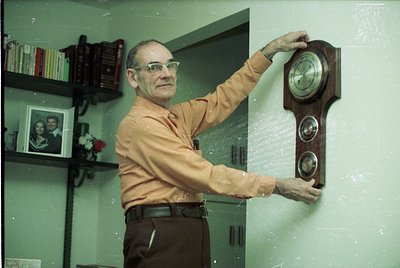 Mid-century office setting featuring a man in a light-colored button-down shirt adjusting a vintage wooden wall clock with du...