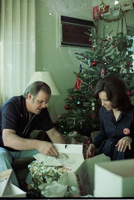 Mid-20th century (likely 1960s–1970s) indoor scene: A man and woman sit on a couch beside a decorated Christmas tree with red...