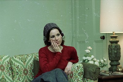 Vintage portrait of a woman in contemplative pose, seated on a floral-patterned armchair against a faded mint-green wall. She...