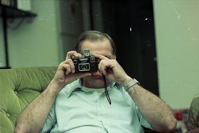Mid-century man holds a vintage **Zeiss Ikon Contax S** camera, framed by a green armchair and muted indoor lighting. Likely ...