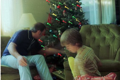 Vintage indoor scene: man and child examining wrapped gifts under a decorated Christmas tree with multicolored lights. 1970s-...