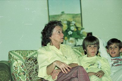 Vintage indoor portrait featuring an elderly woman and two young boys on a floral-patterned sofa. The woman wears a light-col...