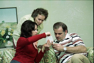 Vintage indoor scene: three adults in mid-1970s clothing (women in long-sleeve blouses, men in striped shirt) gathered around...