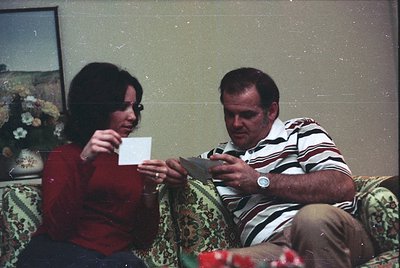 Vintage indoor portrait of two people playing cards on a floral-patterned sofa, 1970s-80s. Woman in red sweater holds a white...