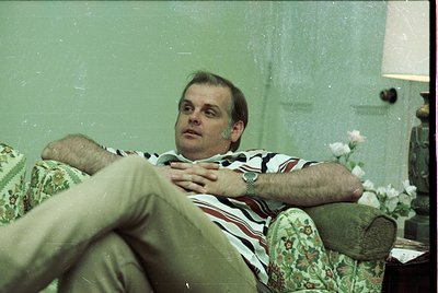 Vintage indoor portrait of a man reclining on a floral-patterned armchair, wearing a striped polo shirt and khaki pants. Soft...