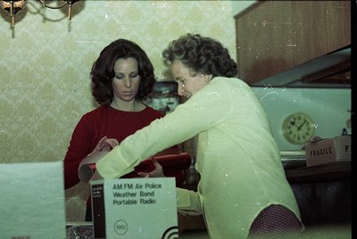 Vintage indoor scene featuring two women examining a vintage **AM/FM Air Police Weather Band Portable Radio** (Tandy brand) i...