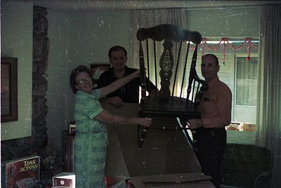 Vintage indoor scene featuring three adults posing with a wooden rocking chair, likely from the 1970s–1980s. Woman in pattern...