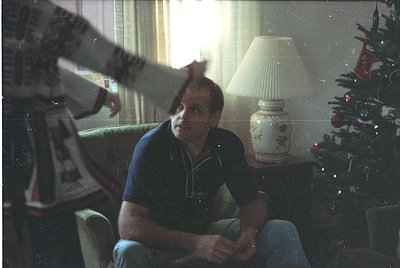 Vintage indoor portrait of a man seated on a mid-century armchair, holding a cigarette. Decor includes a decorated Christmas ...