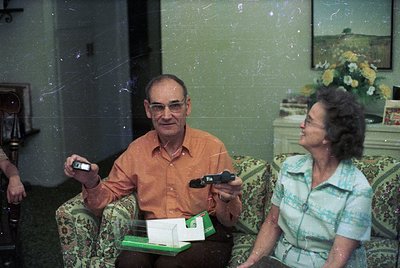 Vintage indoor portrait of two elderly adults in mid-20th-century clothing, likely 1970s–1980s. Man in an orange shirt holds ...