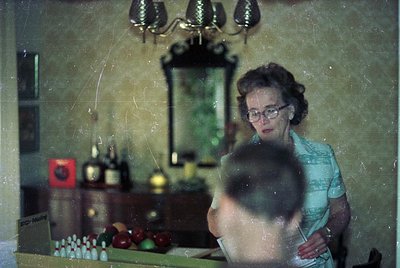 Vintage indoor scene featuring a woman in a light blue blouse standing beside a kitchen countertop adorned with glass bottles...